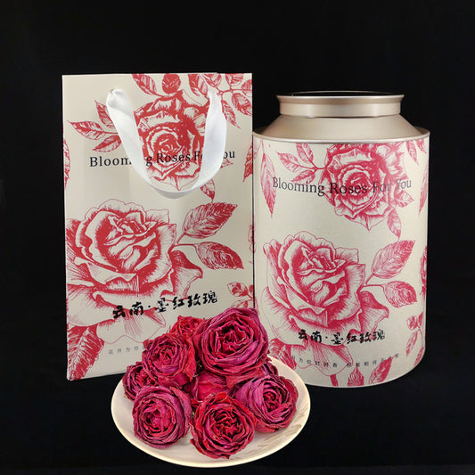 Whole dark red rose crown tea packaging experience featuring the matte white structural sanctuary tin and matching artistic line-art gift carrier bag, fronted by a porcelain dish of whole freeze-dried rose blossoms.